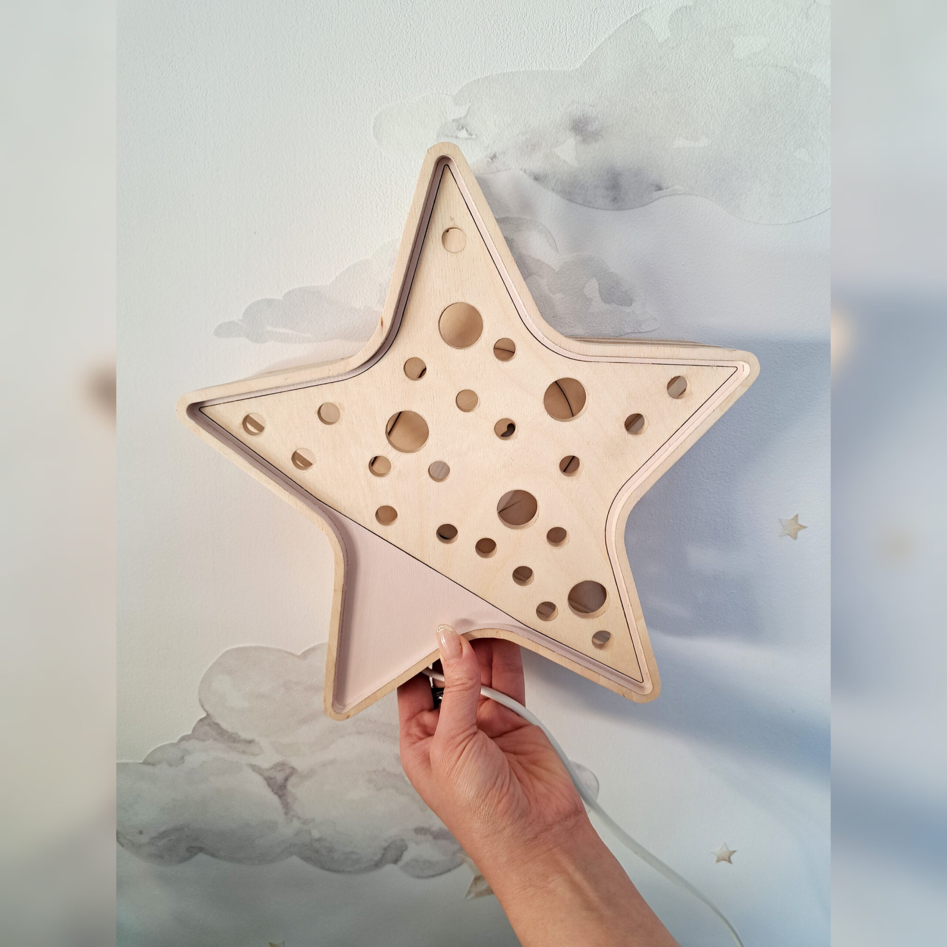 Luminária Estrela de Madeira Controlado por SMARTPHONE Candeeiro de parede em forma de estrela de madeira clara com furos circulares, com mão a segurá-lo e papel de parede de nuvens ao fundo