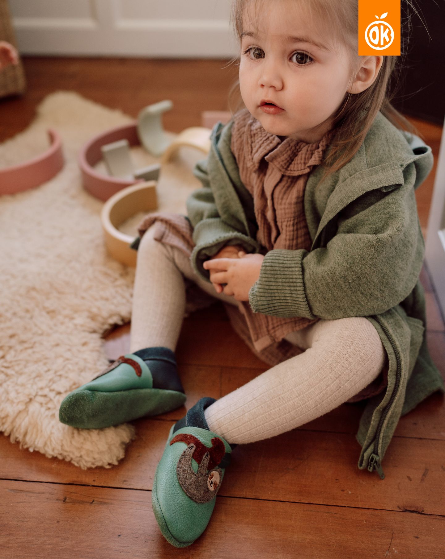 Sapatinhos de Criança Pauli, a Preguiça Bebé com sapatos verde de pele com aplique, vestido castanho e casaco verde sentada no chão de madeira perto de tapete branco felpudo.