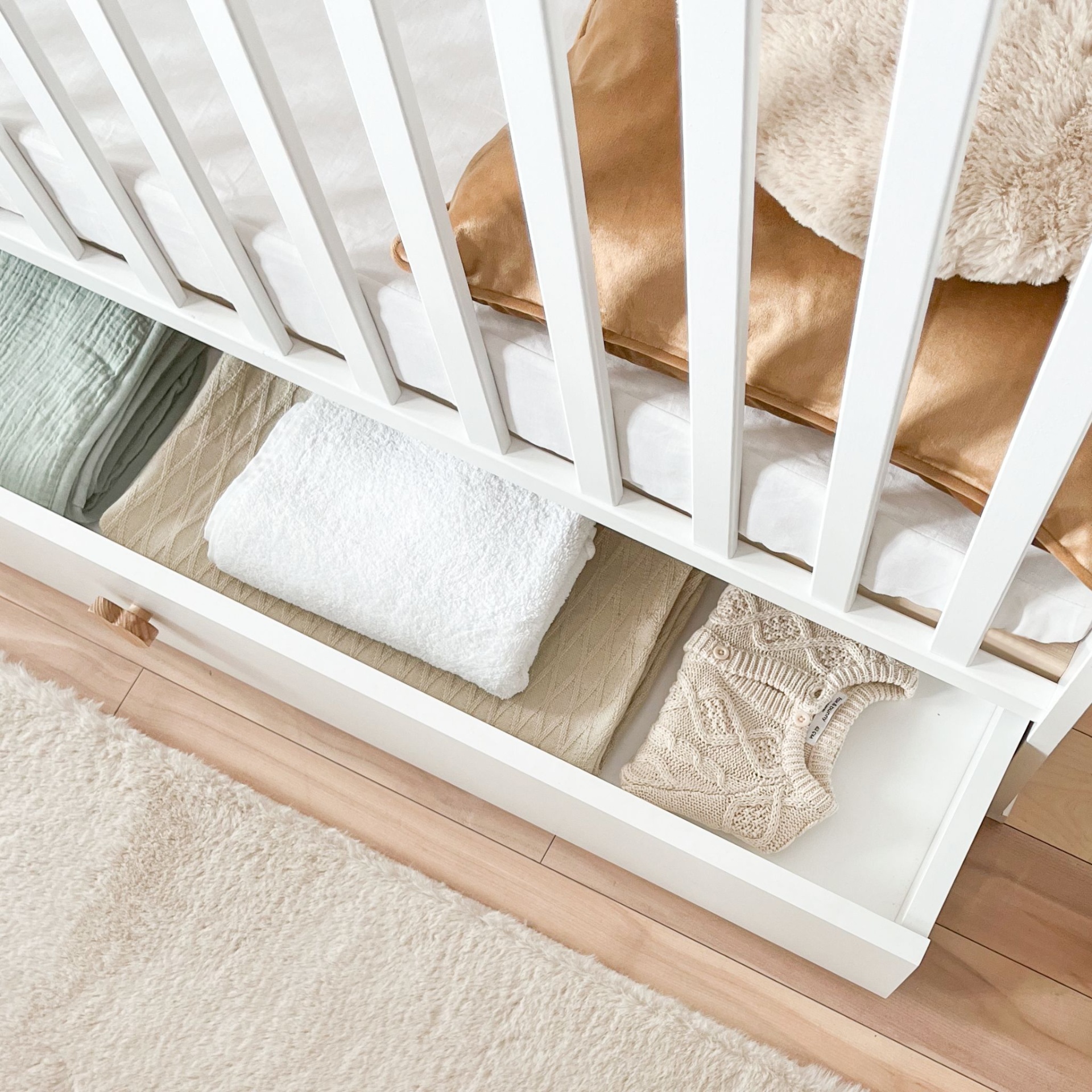 Berço de Bebé PEQUENO CORAÇÃO com Gaveta Berço branco com gaveta aberta mostrando toalha branca, cobertor bege com padrão, malha bege trançada, tecido verde claro, chão madeira clara e tapete creme.