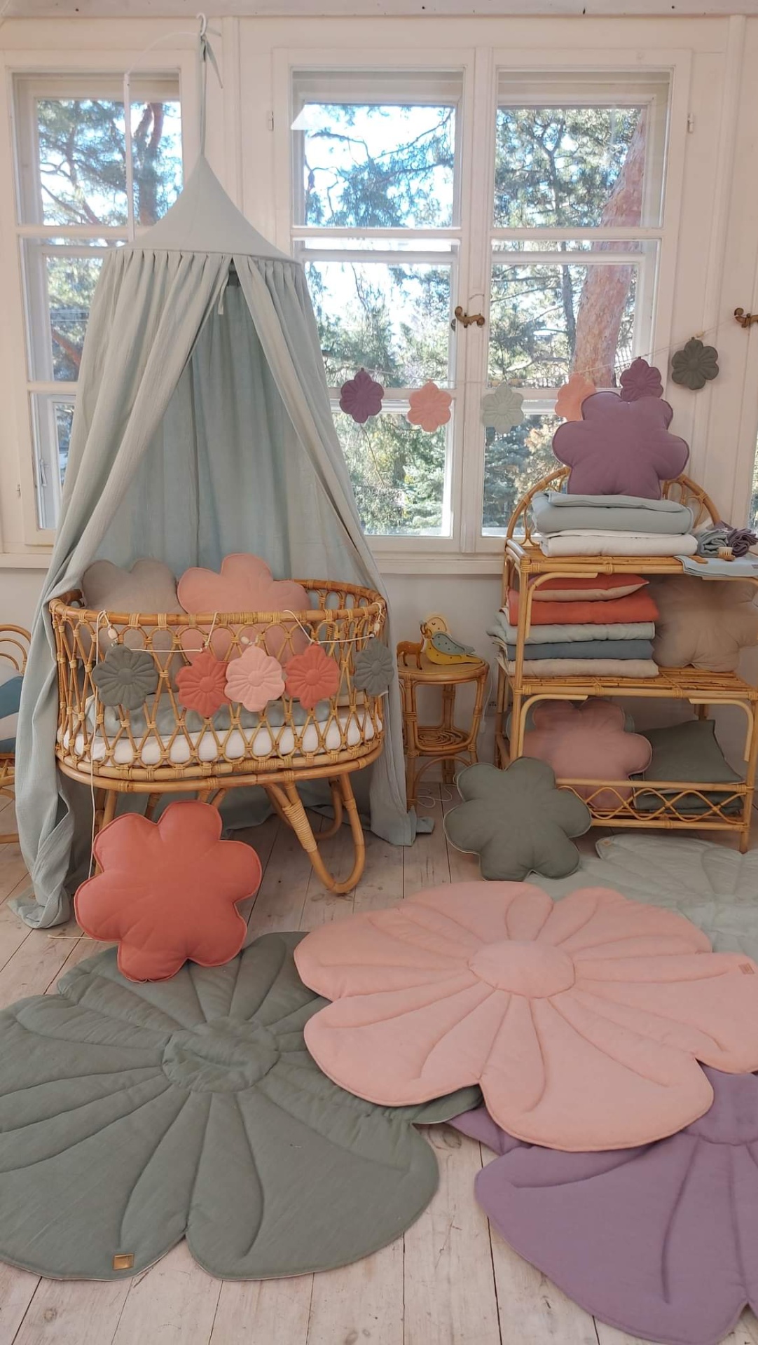 Tapete de Veludo FLOR Berço de bebé em vime com almofadas florais e dossel cinzento num quarto claro com janela e tapetes florais no chão.