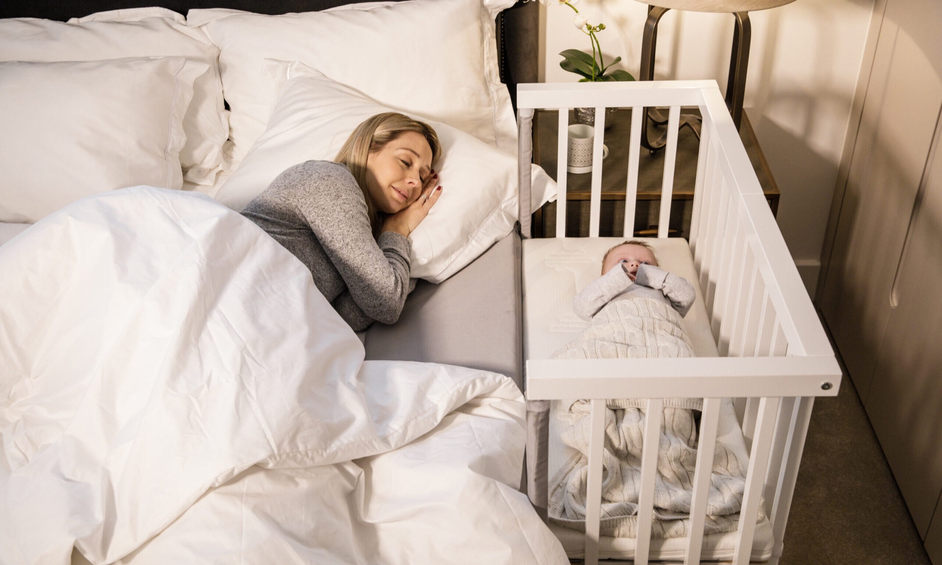 Como Escolher um Berço Co-sleeping para o Bebé e Seus Benefícios Mulher a dormir numa cama ao lado de um berço branco com um bebé enrolado num saco de dormir