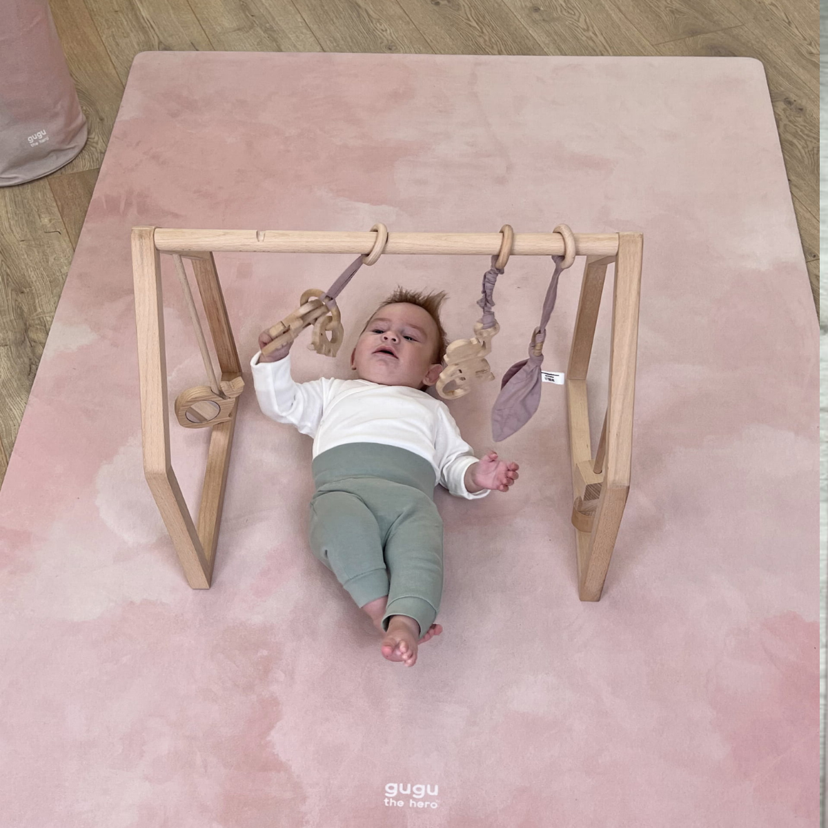 Tapete Grande de Brincar Nuvens Rosa PRIME Ginásio de atividades para bebé em madeira clara com brinquedos e tapete rosa claro com texto 'guau fine baby'.