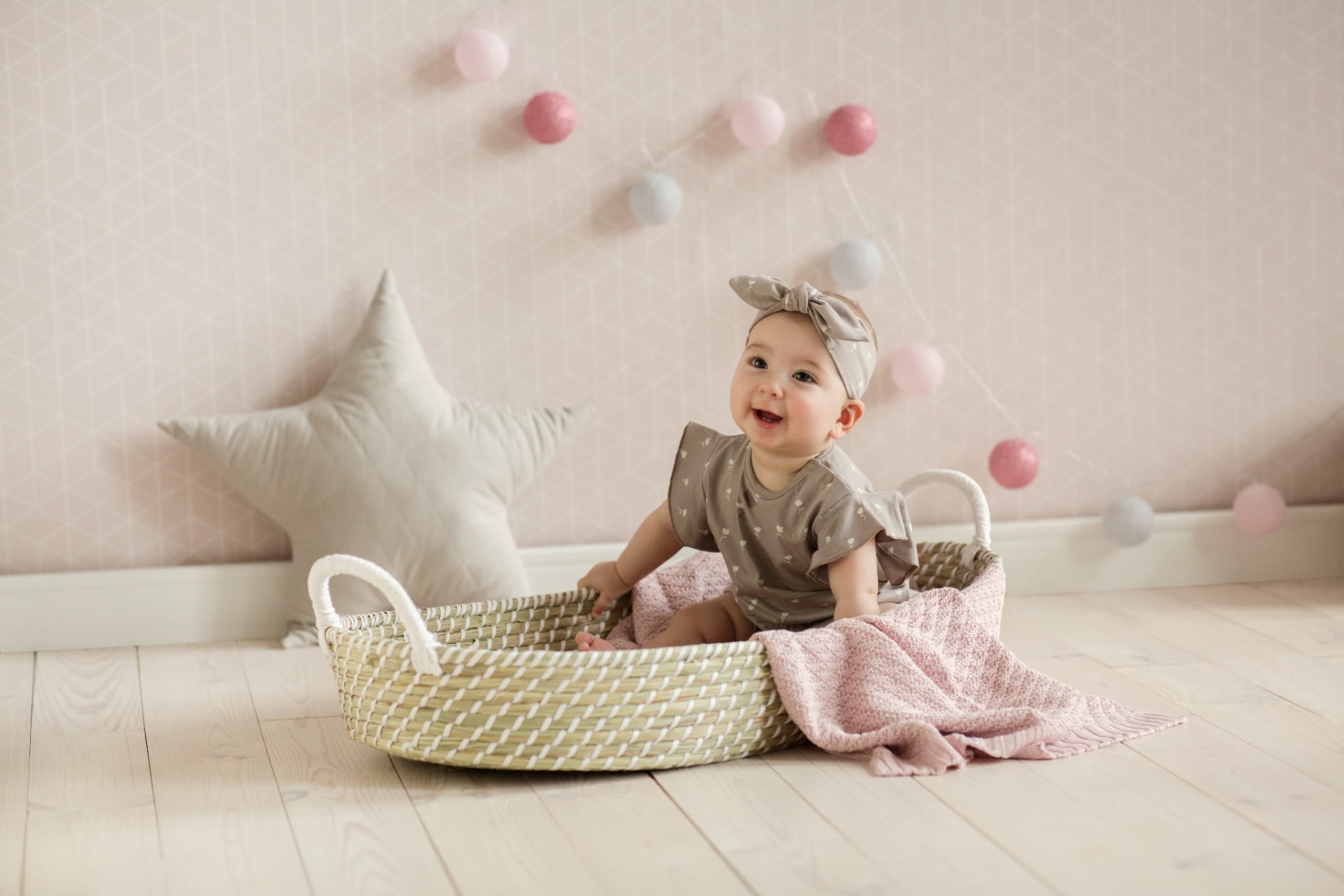 Cesto de Moisés Fraldário em Algas Marinhas Naturais Bebé numa cesta de verga com manta cor-de-rosa e almofada estrela numa sala decorada com guirlanda de bolas