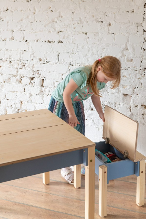 Conjunto de Mesa e Cadeira Infantil com Arrumação Criança ruiva abre caixa de madeira azul clara ao lado de mesa de madeira