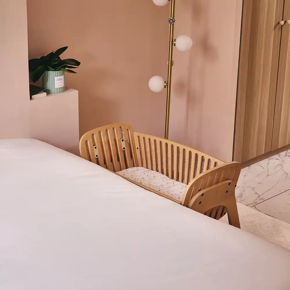 Berço Co-Sleeping KODO em Madeira Natural Com Colchão Berço de madeira clara junto à cama num quarto decorado com parede rosa, candeeiro dourado e planta em vaso verde