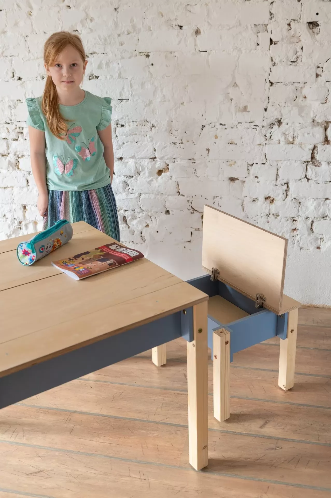 Conjunto de Mesa e Cadeira Infantil com Arrumação Criança ao lado de escrivaninha e cadeira de madeira em interior com parede de tijolos
