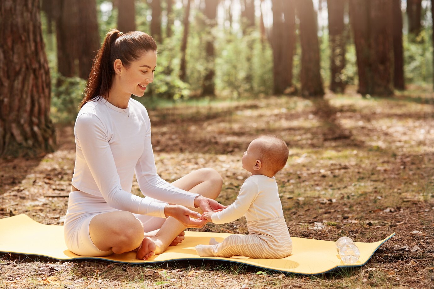 Mulher e bebé vestidos de branco sentados num tapete amarelo de yoga numa floresta