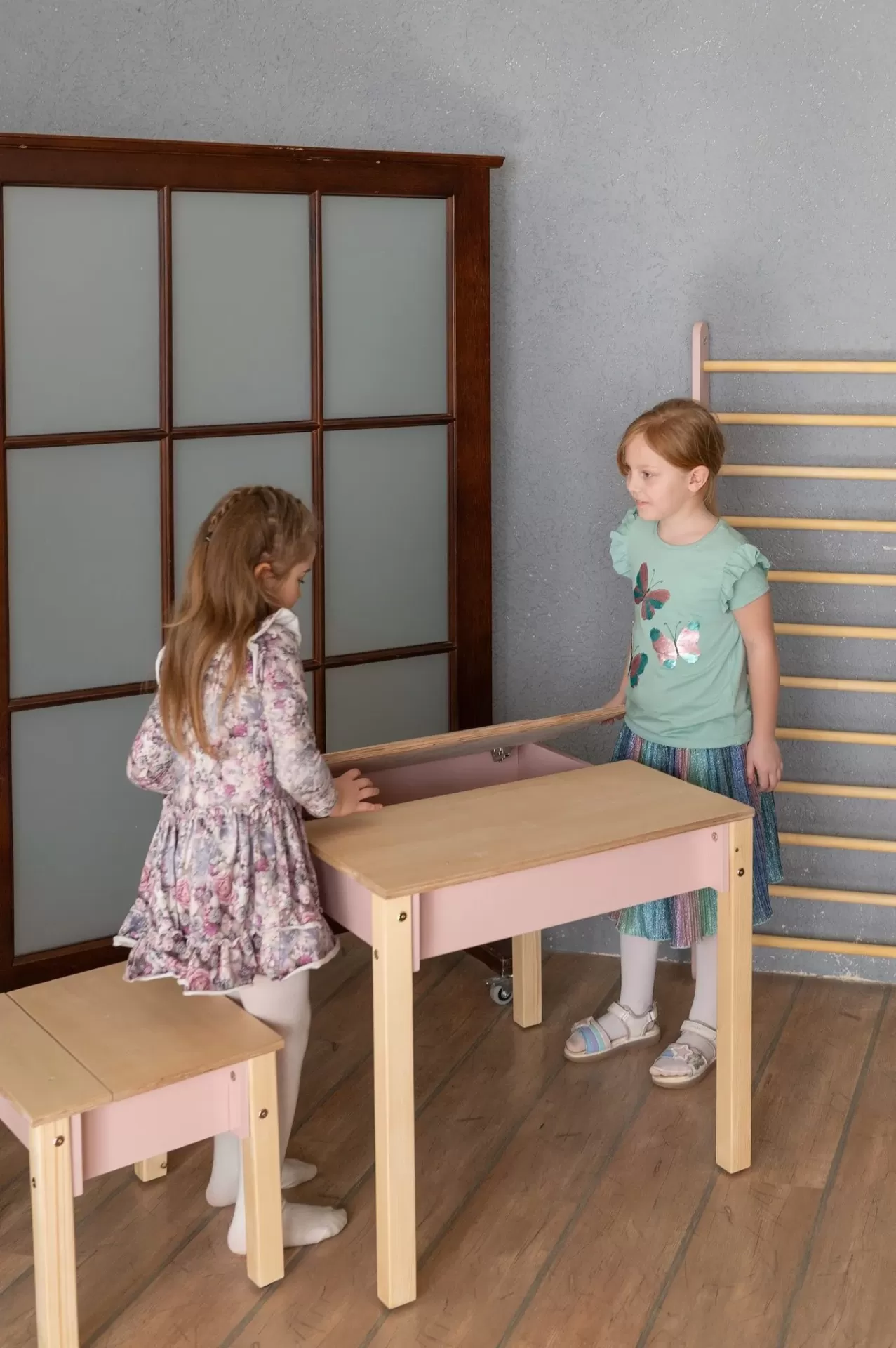 Conjunto de Mesa e Cadeira Infantil com Arrumação Duas meninas a brincar com mesa e bancos infantis de madeira clara e rosa num quarto com parede cinza.