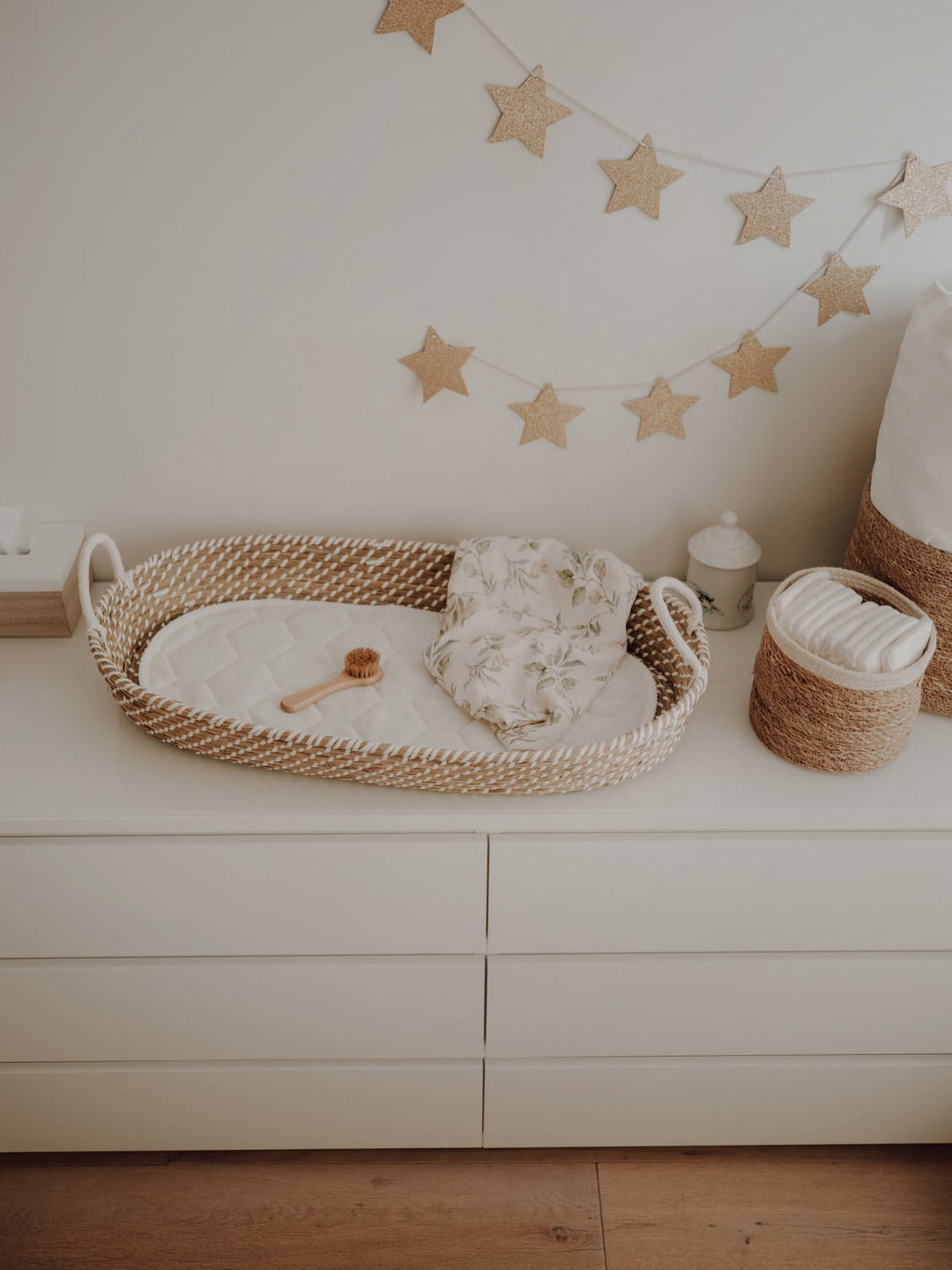 Cesto de Moisés Fraldário em Algas Marinhas Naturais Cesto de vime com cobertores e acessórios de bebê sobre uma cômoda branca, com decoração de estrelas penduradas na parede.