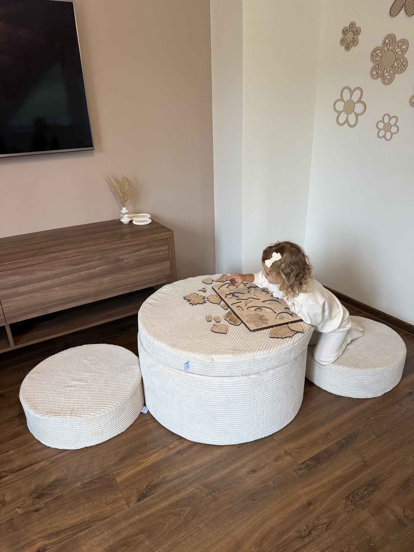 Conjunto Pufe Modular em Veludo Cotelê Bege Mesa redonda e pufes bege em sala com chão de madeira e parede decorada, criança brinca com jogo de peças de madeira