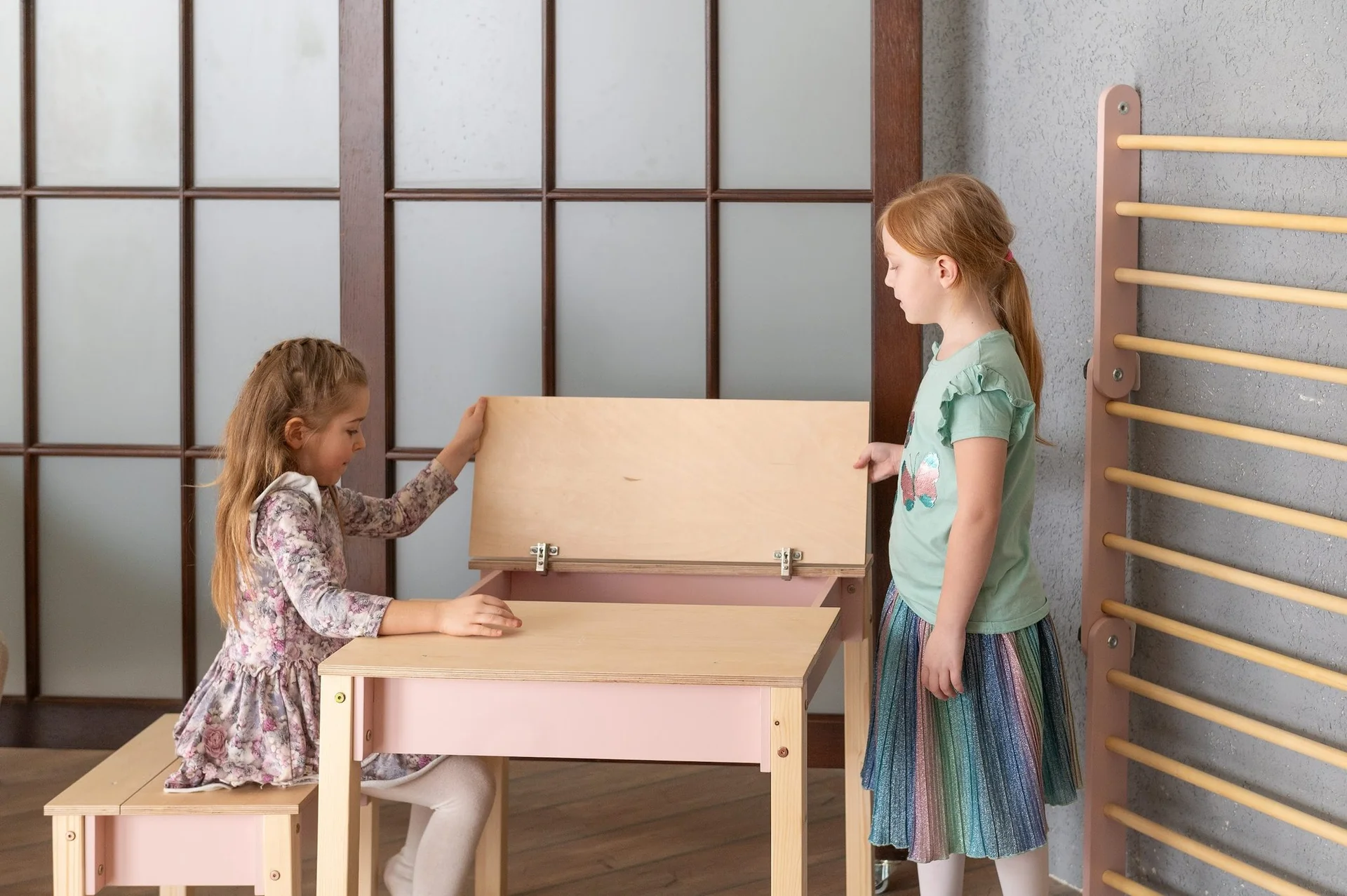 Conjunto de Mesa e Cadeira Infantil com Arrumação Mesa e banco infantil em madeira clara e rosa com tampo articulado e duas crianças num interior com porta de vidro e parede cinza