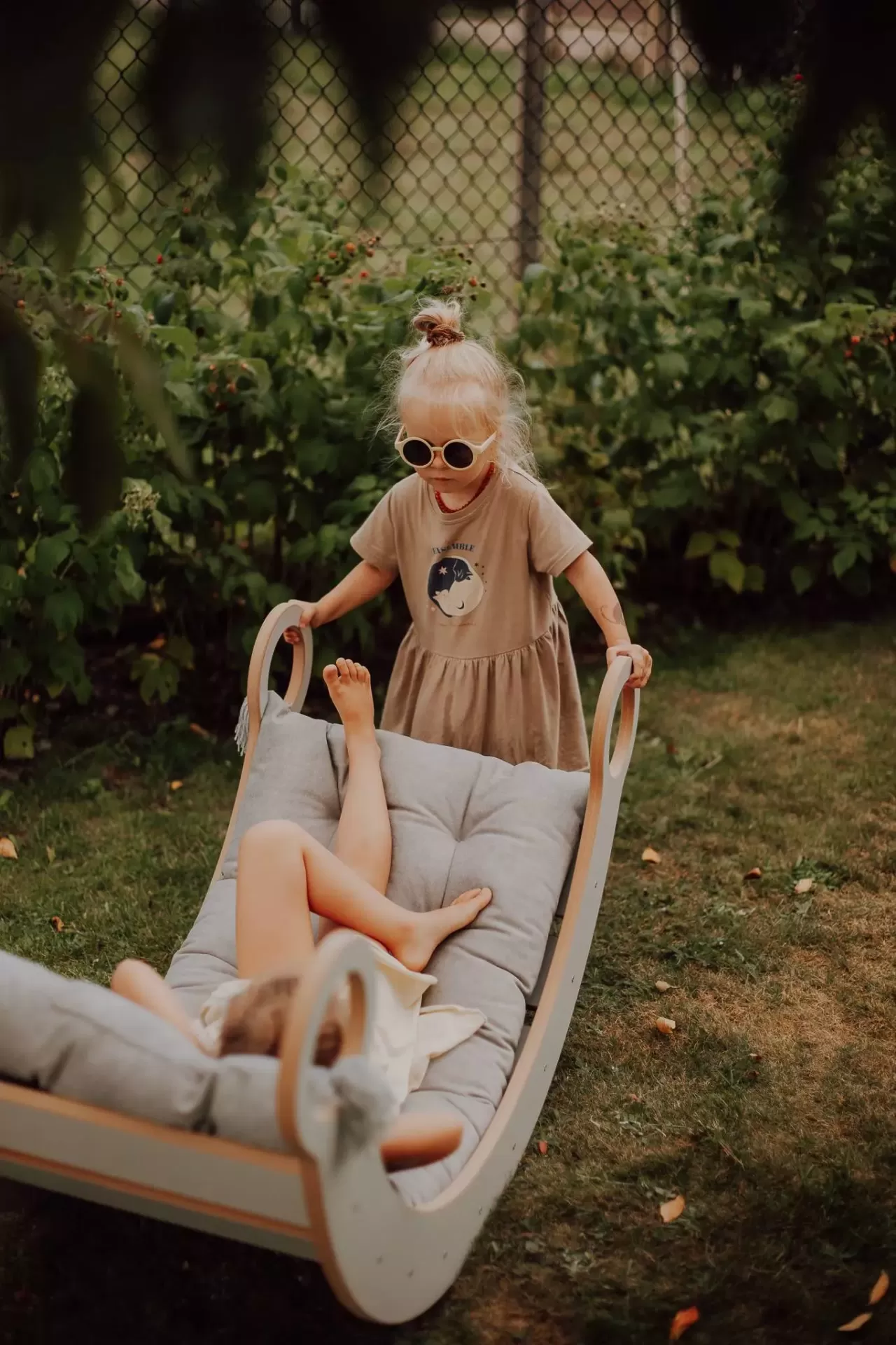 Balanço de Madeira MONTESSORI Duas crianças a brincar numa cadeira balanço de madeira com almofadas cinzentas num jardim