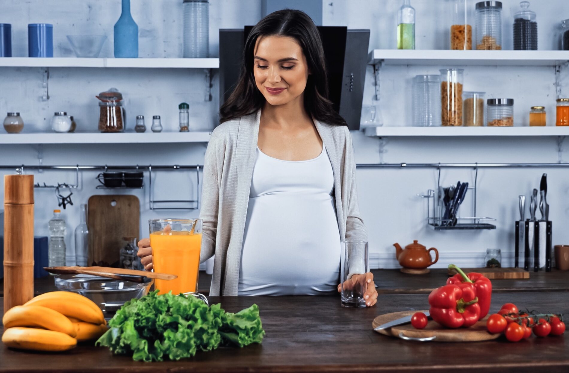 Mulher grávida em cozinha moderna com frutas e vegetais sobre bancada de madeira.