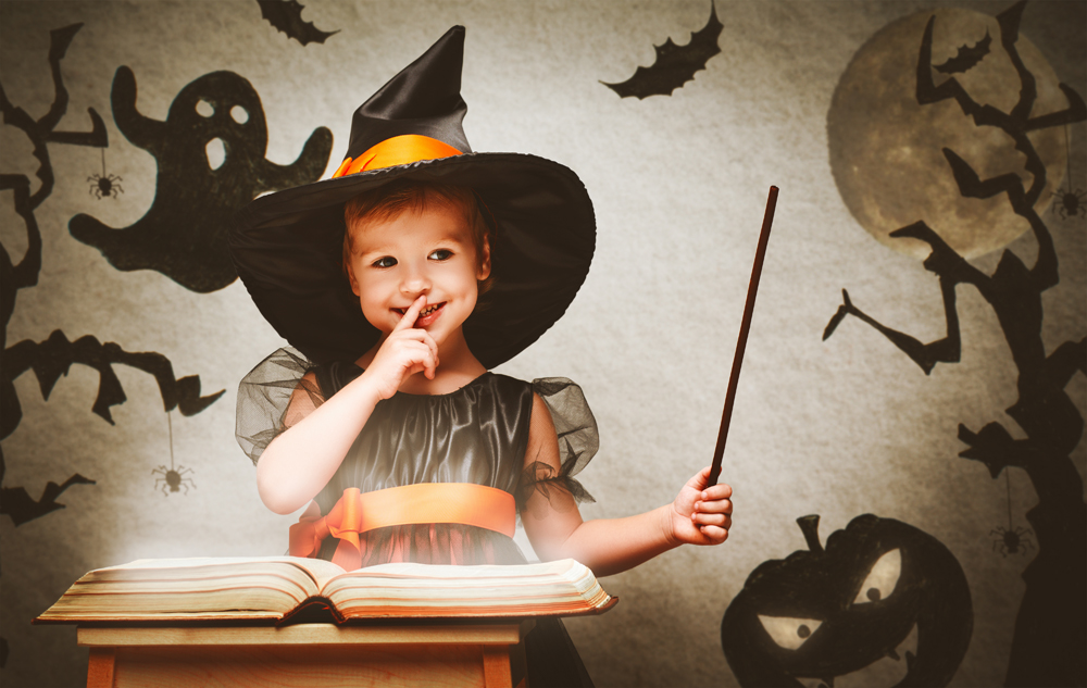 Criança vestida de bruxa segurando varinha com livro aberto e fundo decorado de Halloween