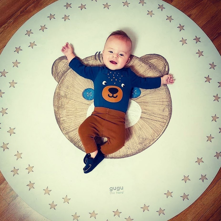 Tapete Redondo Borracha Natural URSO TEDDY Bebé deitado num tapete redondo branco com estrelas castanhas e rosto de urso no centro
