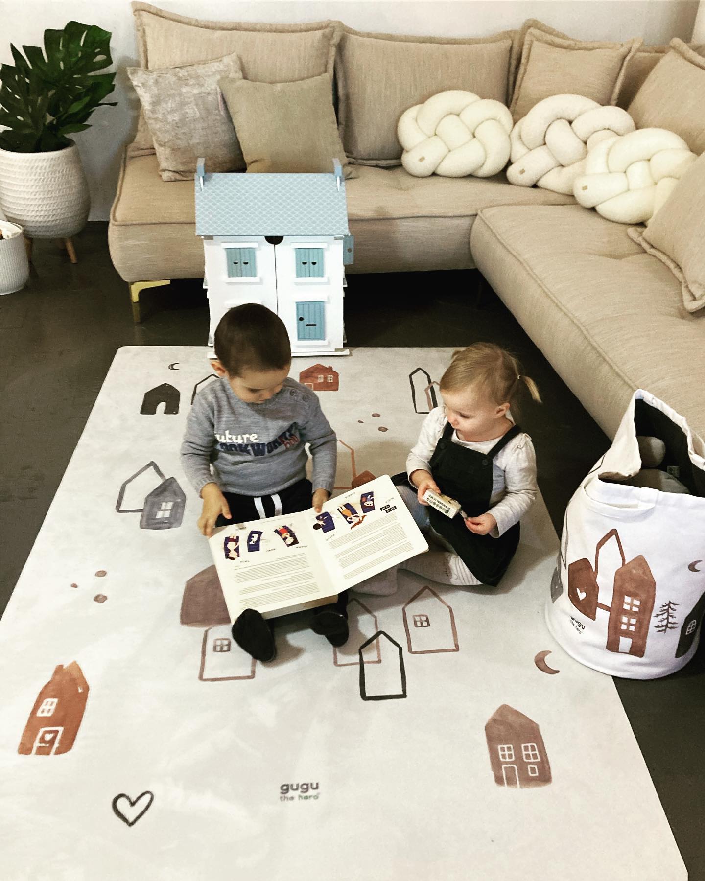 Tapete Retangular Ecológico CIDADE ESCANDINAVA Tapete infantil branco com casas castanho, cinza e preto, crianças sentadas a ler, sofá bege e casa de brinquedo branca com detalhes azuis