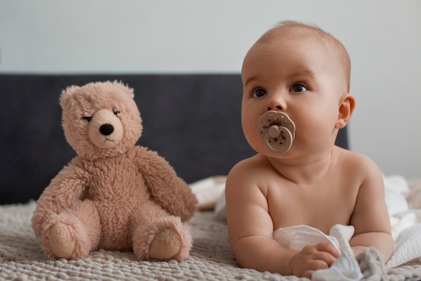 Bebé com chupeta castanha e urso de pelúcia castanho claro ao lado numa manta bege.