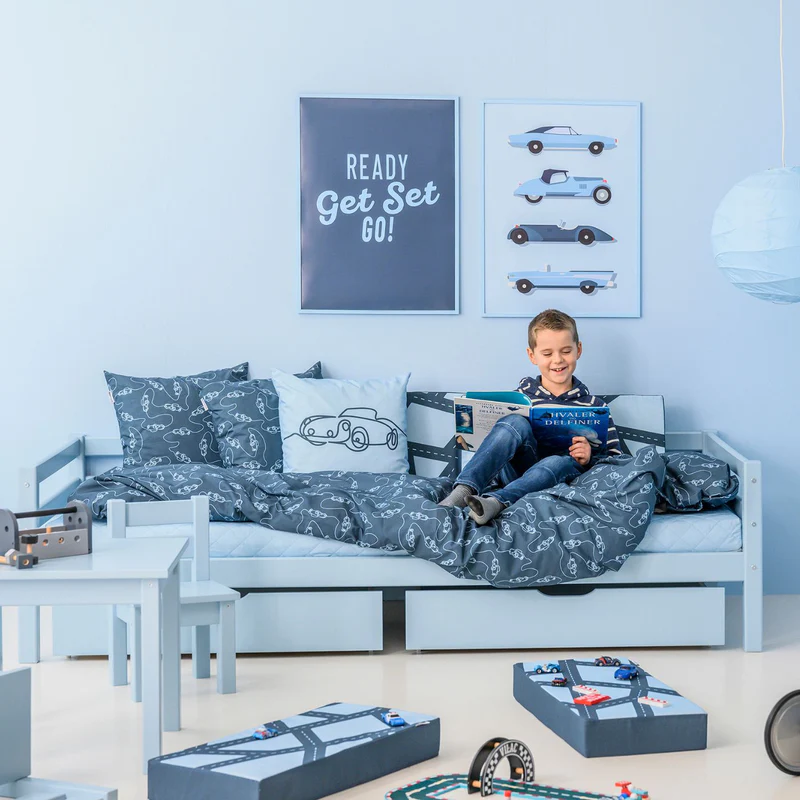 Conjunto de Edredão e Fronha Infantil – Carro Quarto infantil com cama azul claro, roupa de cama cinzenta com padrão de carros, almofada branca com desenho de carro, quadros na parede com texto e imagens, mesa e cadeiras azuis e brinquedos no chão
