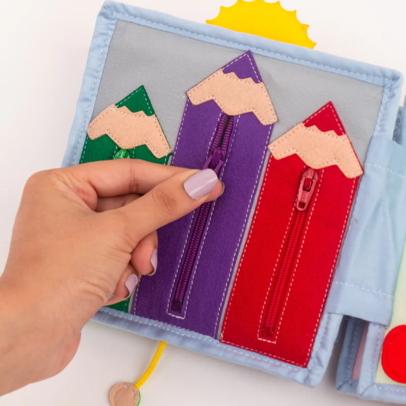 Arco Íris - Mini Livro Sensorial de 6 Páginas Livro educativo de tecido com lápis coloridos e fechos de correr sendo aberto por uma mão com unhas lilás.