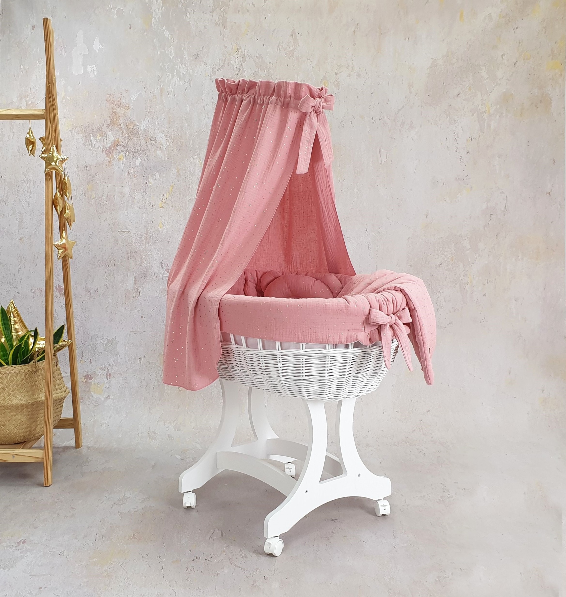 Cesta Moisés de Vime Branco EMMA com Roupa de Cama Berço oval branco com suporte branco e têxtil rosa com dossel num ambiente neutro com estante de madeira e plantas
