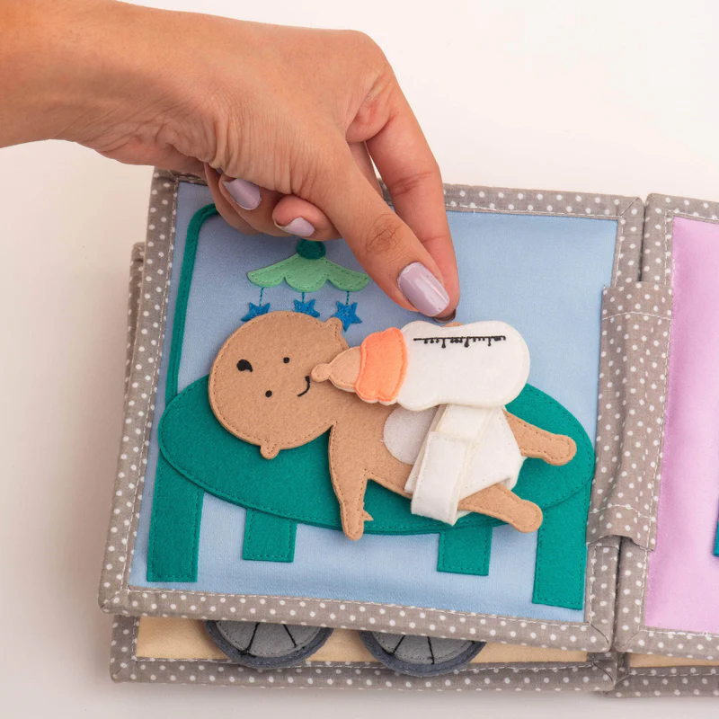 Bebé Feliz – MINI Livro Sensorial de 6 Páginas Livro têxtil infantil com figura de bebê a ser alimentado com biberão.