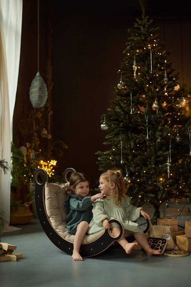 Balanço de Madeira MONTESSORI Duas crianças sentadas num baloiço de madeira perto de árvore de Natal com luzes e presentes.