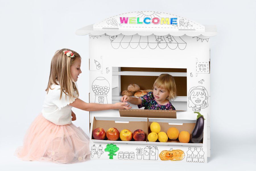 Minimercado para Colorir Duas crianças brincam numa casinha de papelão branca decorada, com frutas de plástico em exposição.