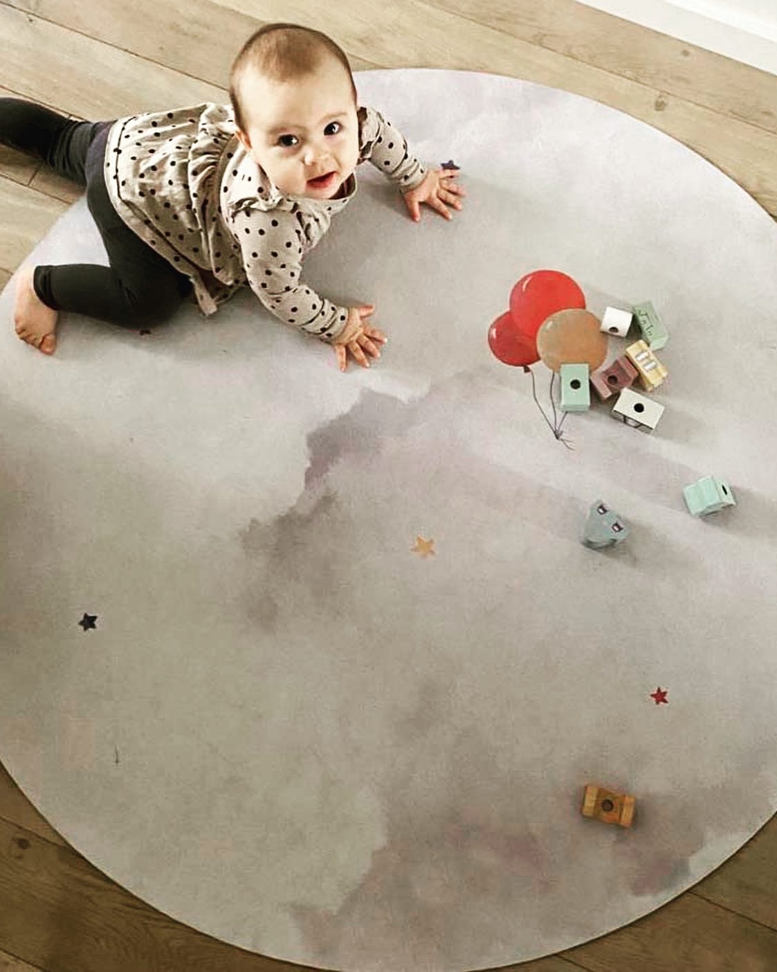 Tapete Redondo de Borracha Natural Balões PRIME Bebé em tapete redondo cinza claro com padrão de nuvens e estrelas pequenas, brinquedos de madeira coloridos, chão de madeira