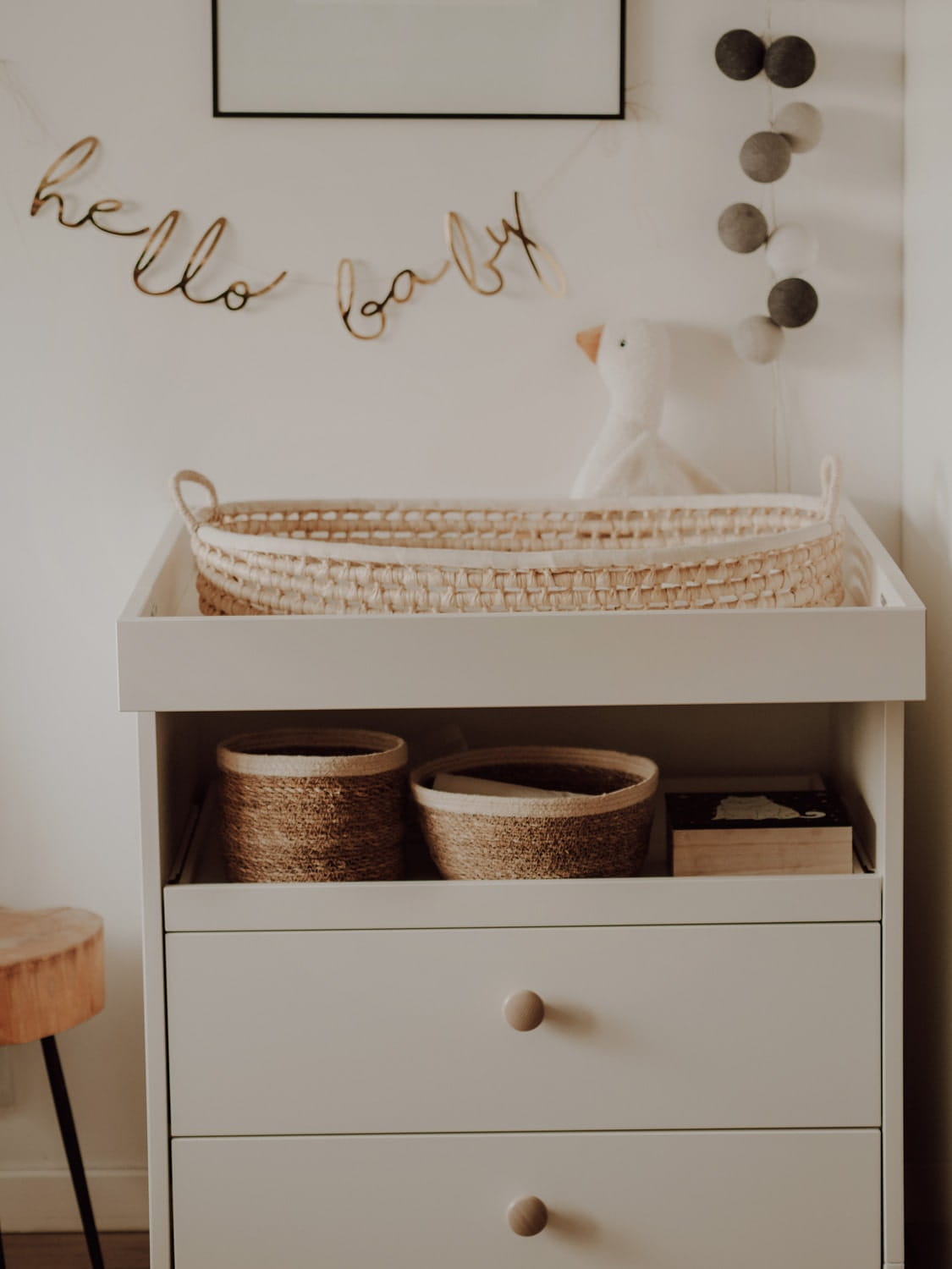 Cesto Fraldário de CASCA de MILHO Móvel branco com gavetas, cestos castanhos, berço de vime, decoração de parede com texto 'hello baby' e pendentes de bolas cinzentas