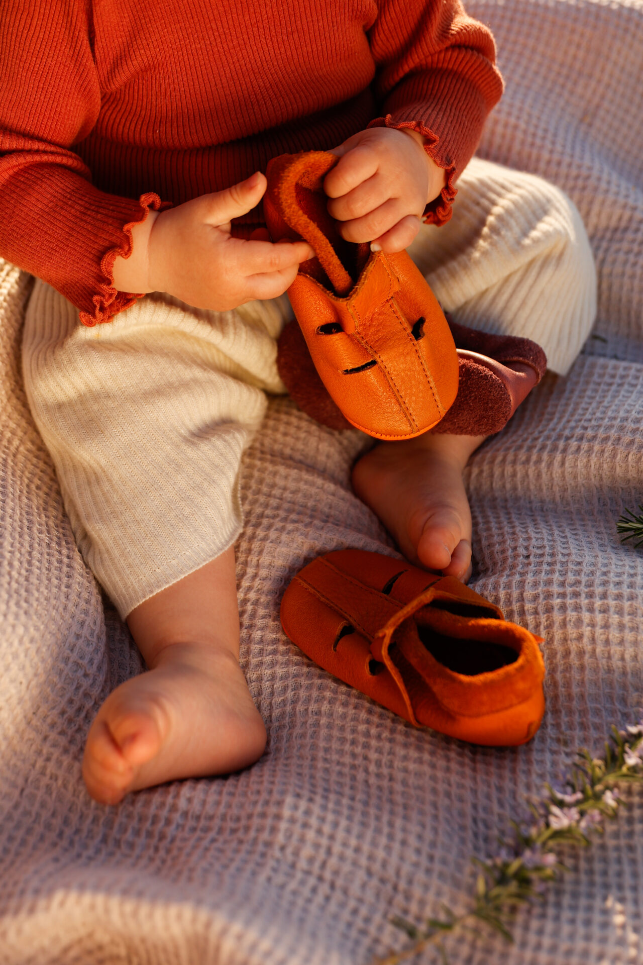 Sandália Infantil Azul Sapatos de pele castanha alaranjada para bebé com detalhe preto, bebé com roupa bege e tijolo sentado