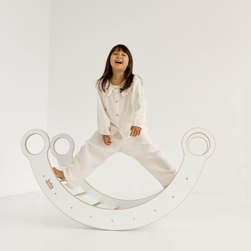 Balanço de Madeira MONTESSORI Criança numa balança de madeira branca num ambiente branco.