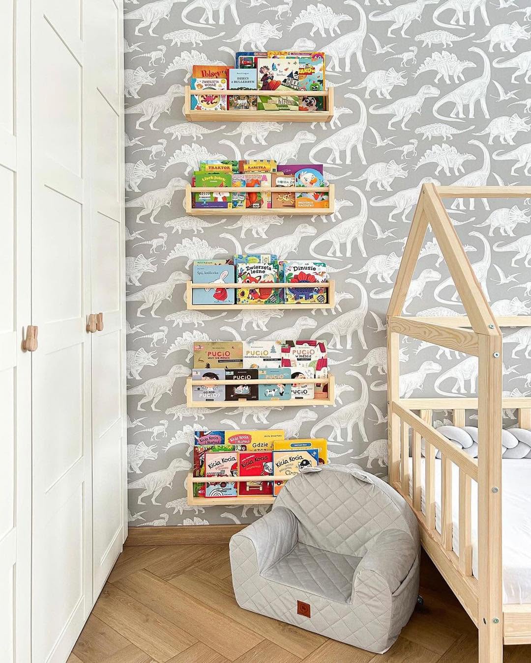 Papel de Parede Infantil Dinossauros Cinza Quarto infantil com papel de parede de dinossauros, estantes de madeira com livros, cadeira pequena cinza e cama de madeira clara.