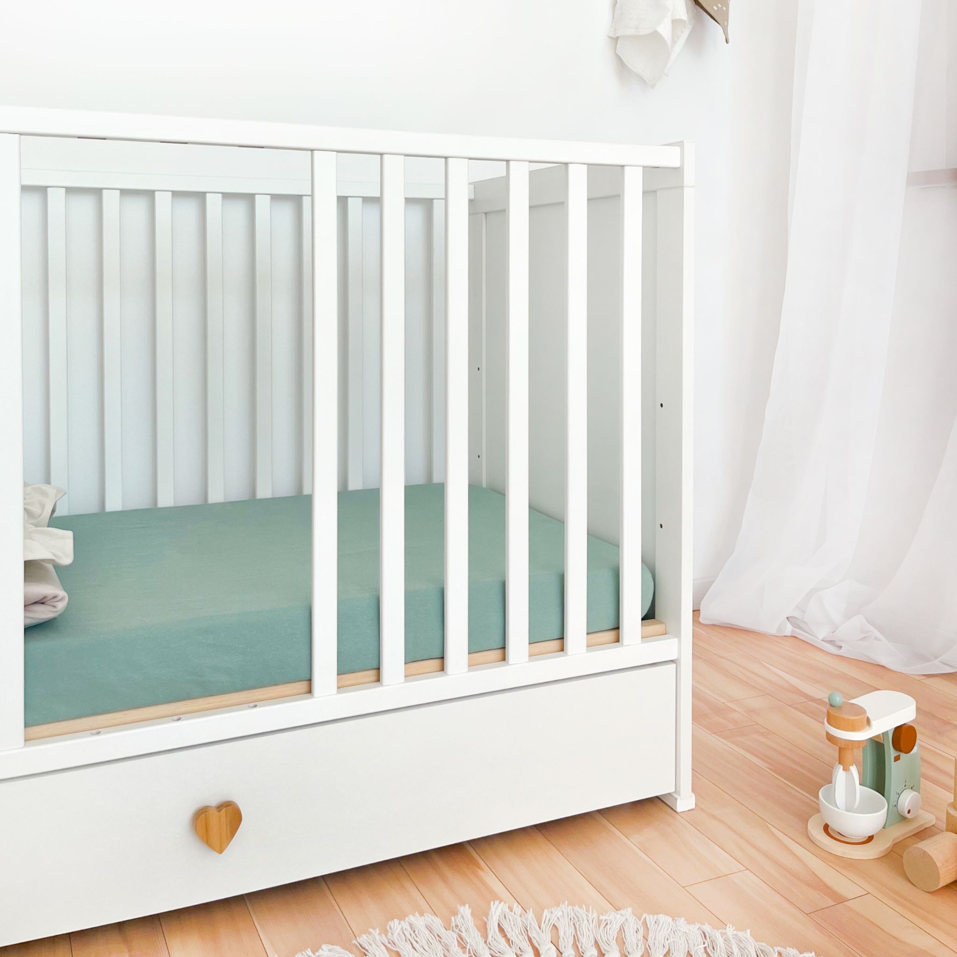 Berço de Bebé PEQUENO CORAÇÃO com Gaveta Berço branco de madeira com colchão azul esverdeado e puxador de coração, com brinquedo de madeira ao lado e cortinas brancas.