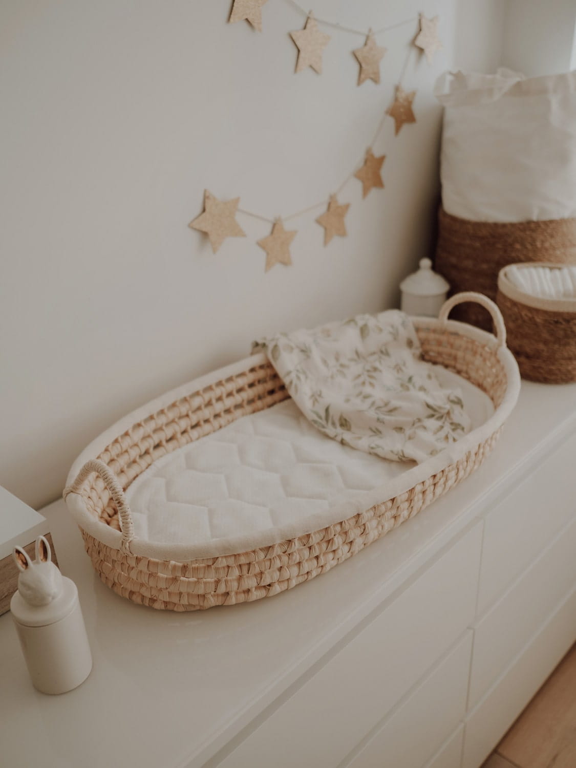 Cesto Fraldário de CASCA de MILHO Berço de vime com colchão branco e manta floral em móvel branco com guirlanda de estrelas na parede