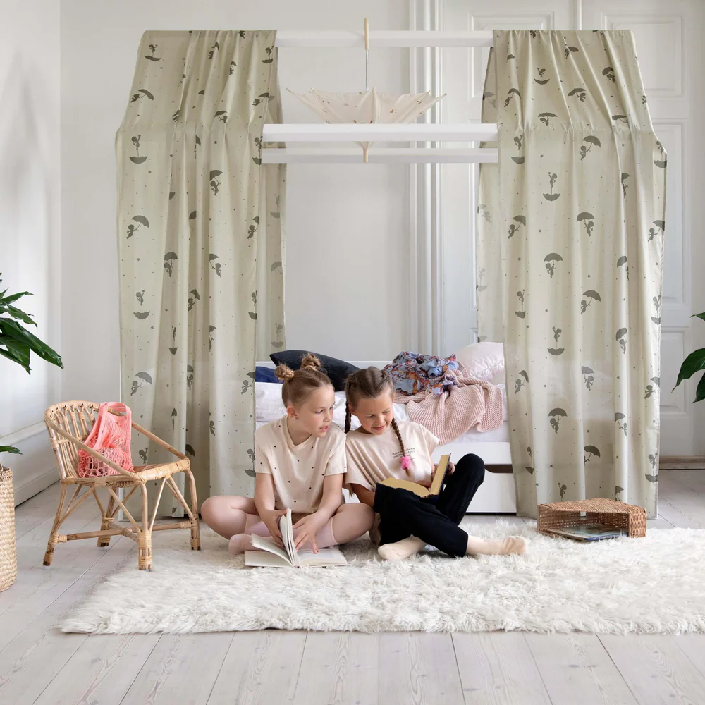 Quarto infantil com cortinas bege com padrão de guarda-chuvas, crianças a ler no tapete branco felpudo.