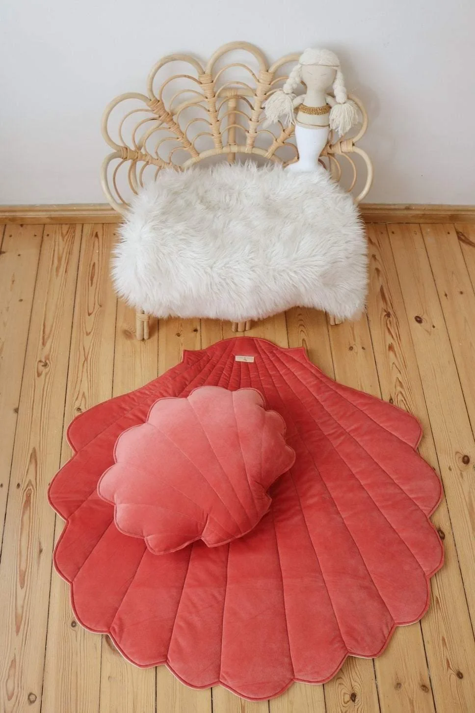 Tapete Veludo CONCHA Tapete e almofada decorativos rosa em chão de madeira clara com cadeira de rattan e pelúcia branca ao fundo