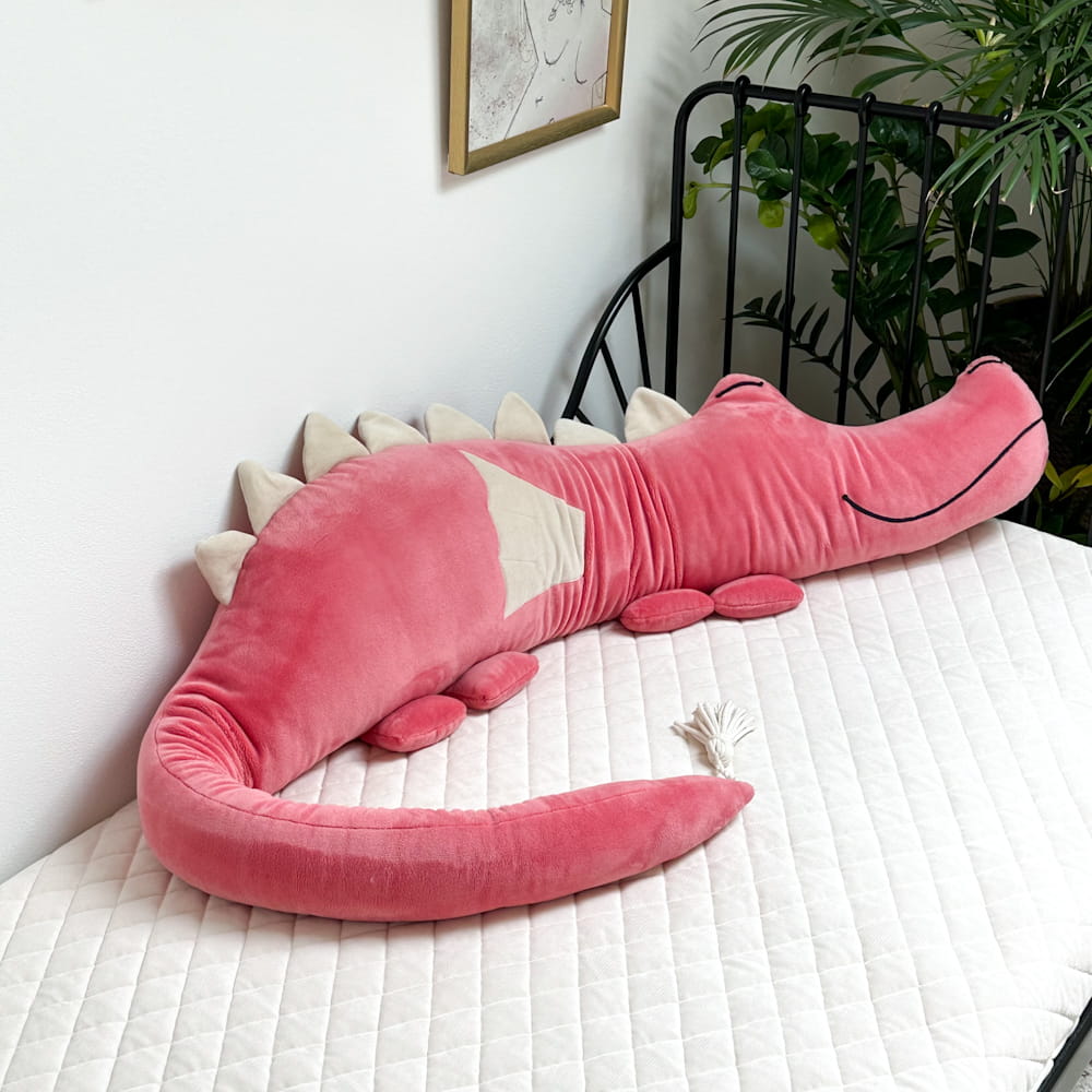 Protetor de Cama/Berço DRAGÃO PELÚCIA Pelúcia de crocodilo cor-de-rosa numa cama branca com cabeceira preta