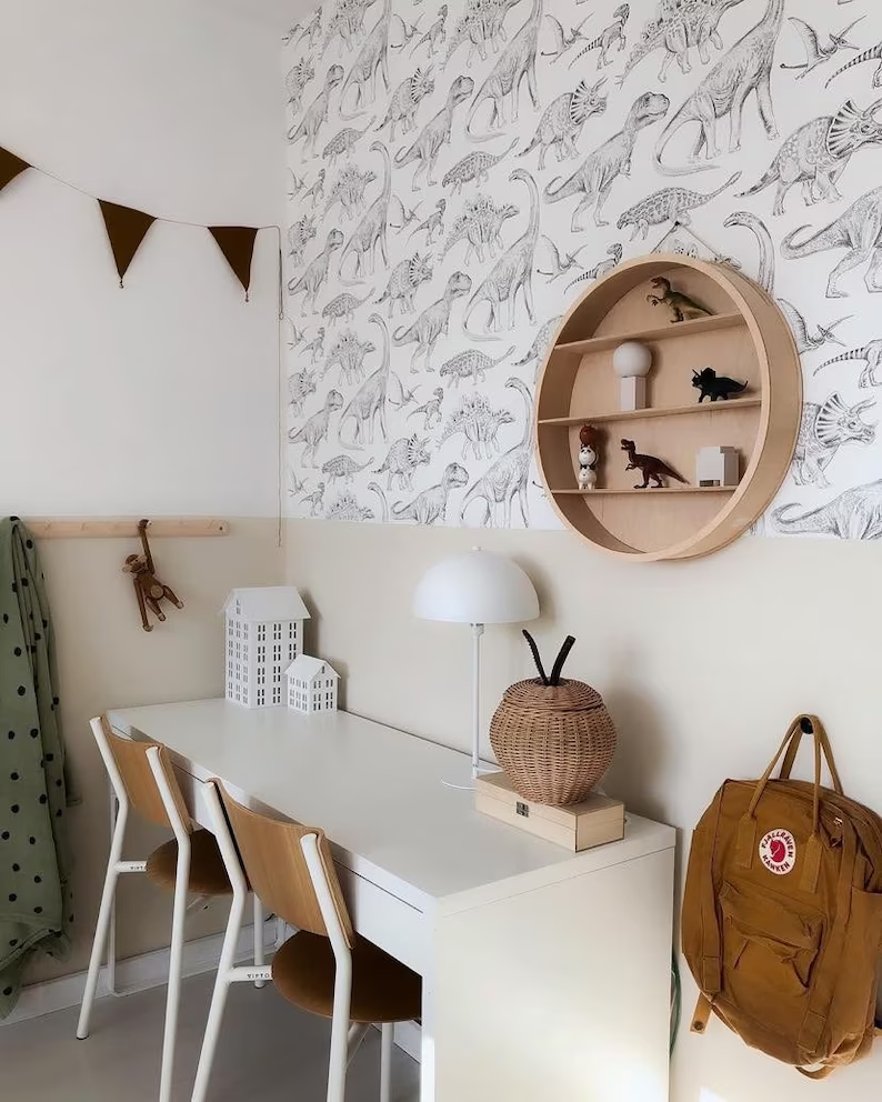 Papel de Parede Dinossauros BRANCO Quarto infantil com secretária, cadeiras, candeeiro, prateleira redonda com dinossauros e papel de parede com padrão de dinossauros.