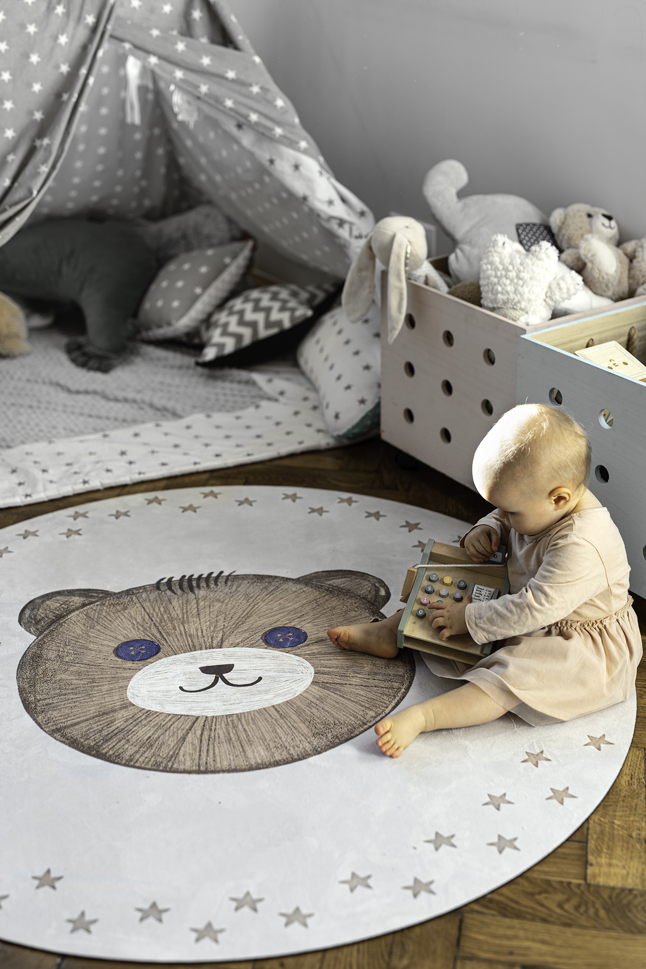 Tapete Redondo Borracha Natural URSO TEDDY Criança sentada num tapete redondo com desenho de urso e estrelas, brincando com brinquedo de madeira, ao lado de caixa de brinquedos e tenda cinzenta com estrelas.