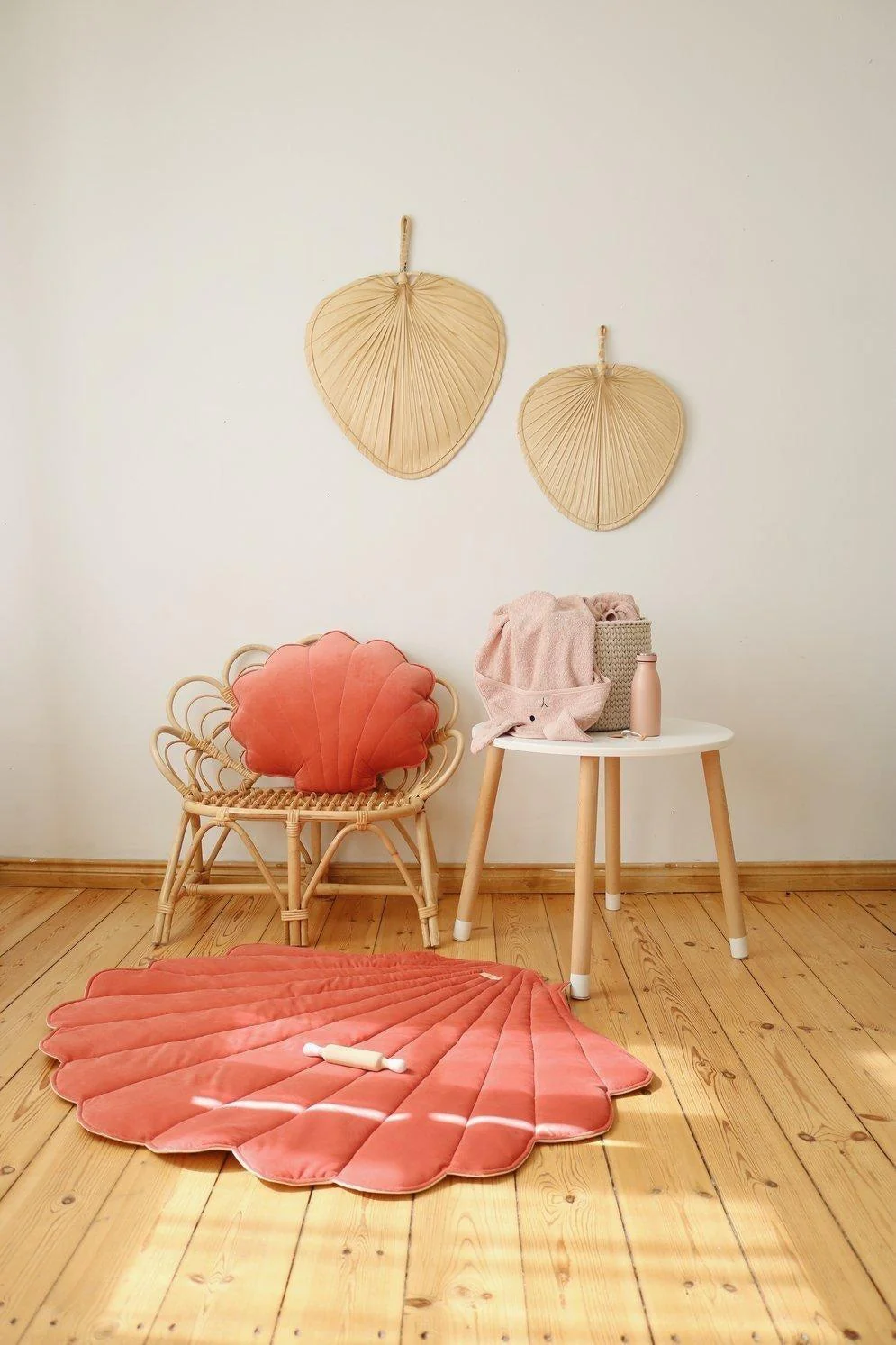 Tapete Veludo CONCHA Tapete e almofada em forma de concha cor-de-rosa, cadeirão de rattan, mesa branca com toalha e recipiente, chão de madeira clara.