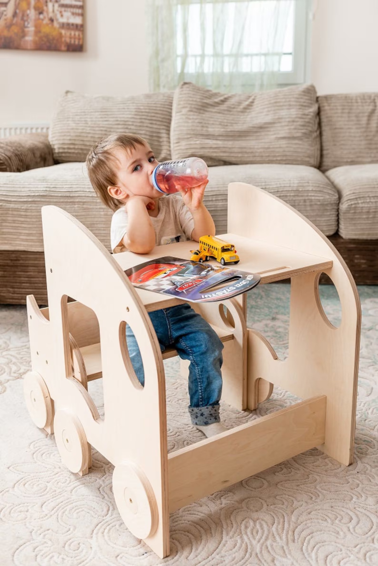 Mesa e Cadeira Infantil MONTESSORI Criança numa secretária de madeira em formato de carrinho com brinquedos e sofá ao fundo
