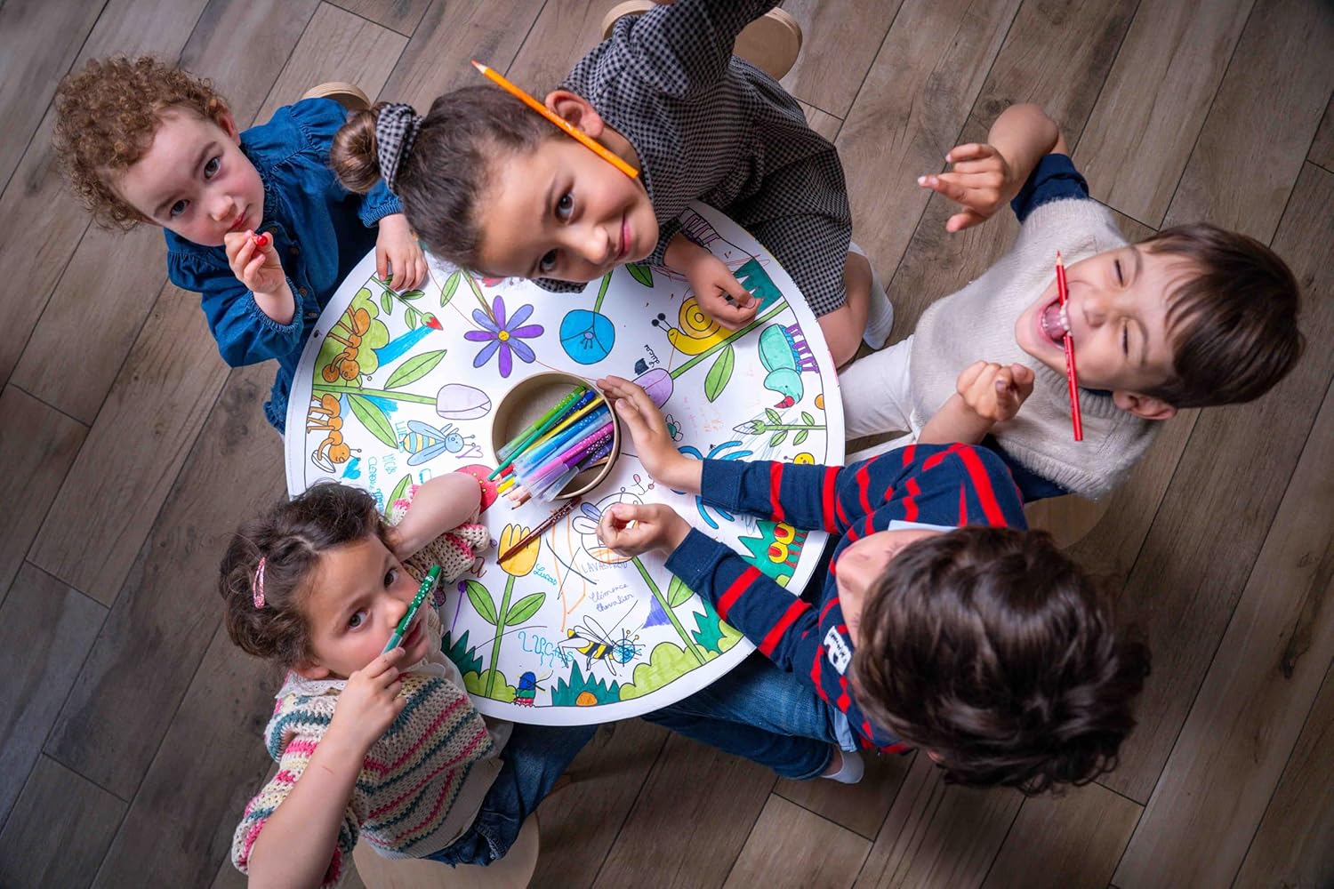 Recarga de Folhas para Colorir - Coelhinhos Mesa redonda com desenhos coloridos e cinco crianças a desenhar com lápis de cor numa sala com chão de madeira.