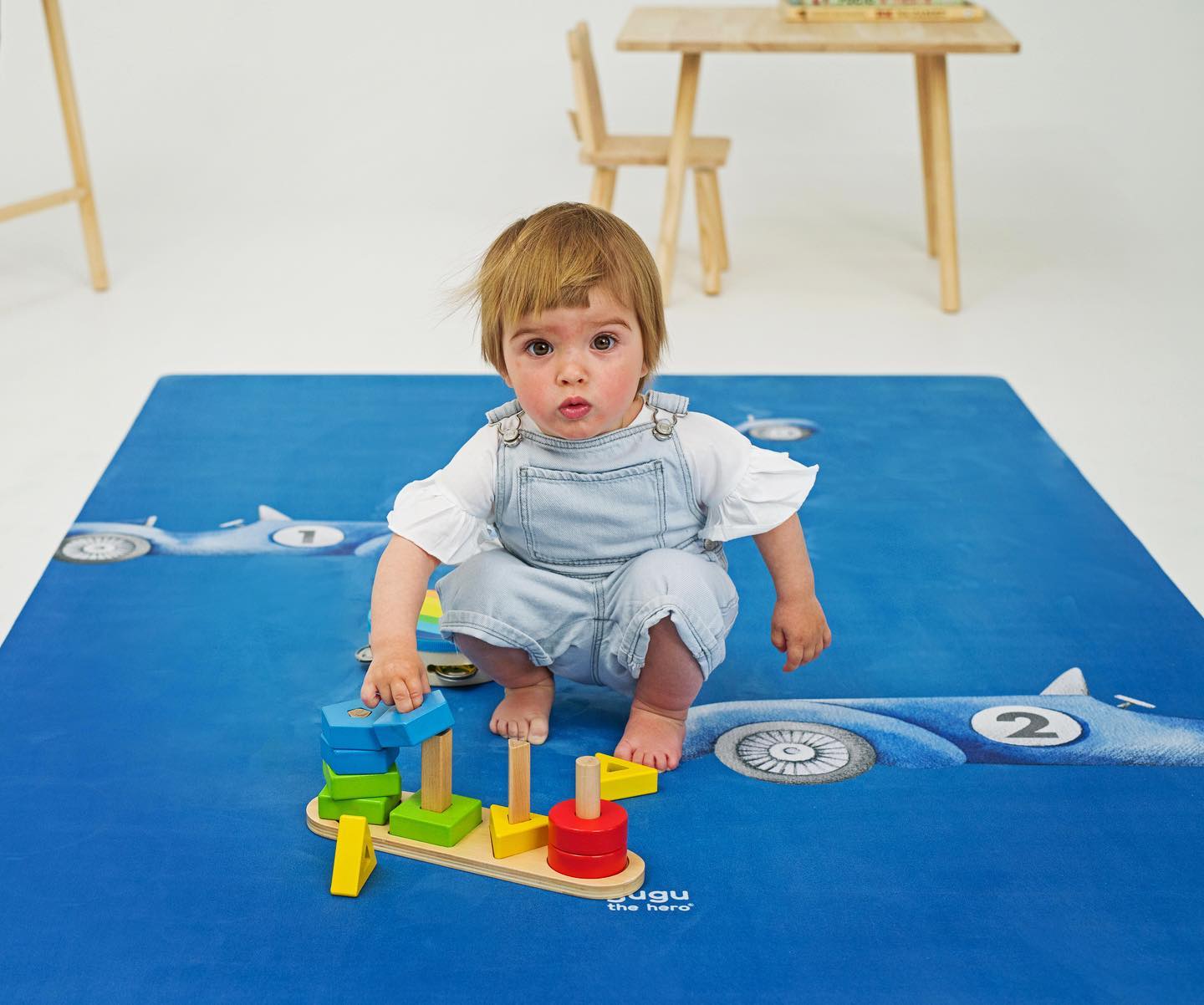 Tapete Educativo ECO Carros de Corrida PRIME Criança sentada a brincar num tapete azul com padrão de aviões e números.