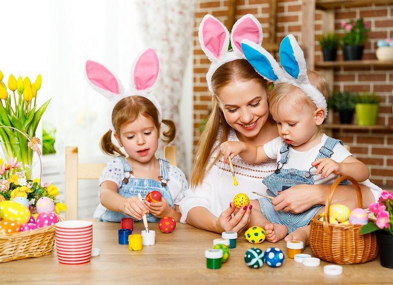 Crianças e mulher pintam ovos de Páscoa com orelhas de coelho na mesa, cestos com ovos e flores ao lado.