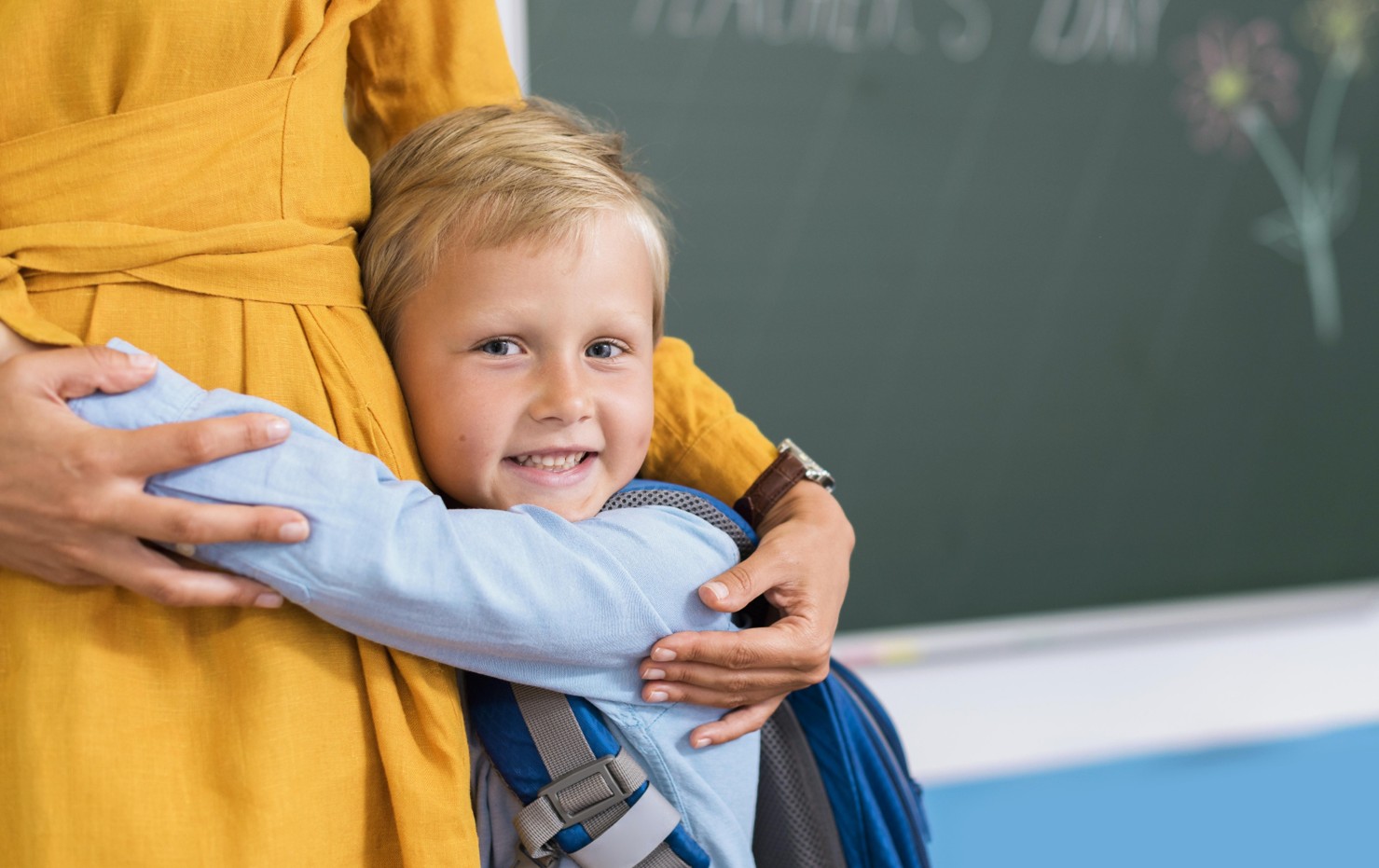 Criança com mochila azul abraçada a adulto com túnica amarela em sala de aula