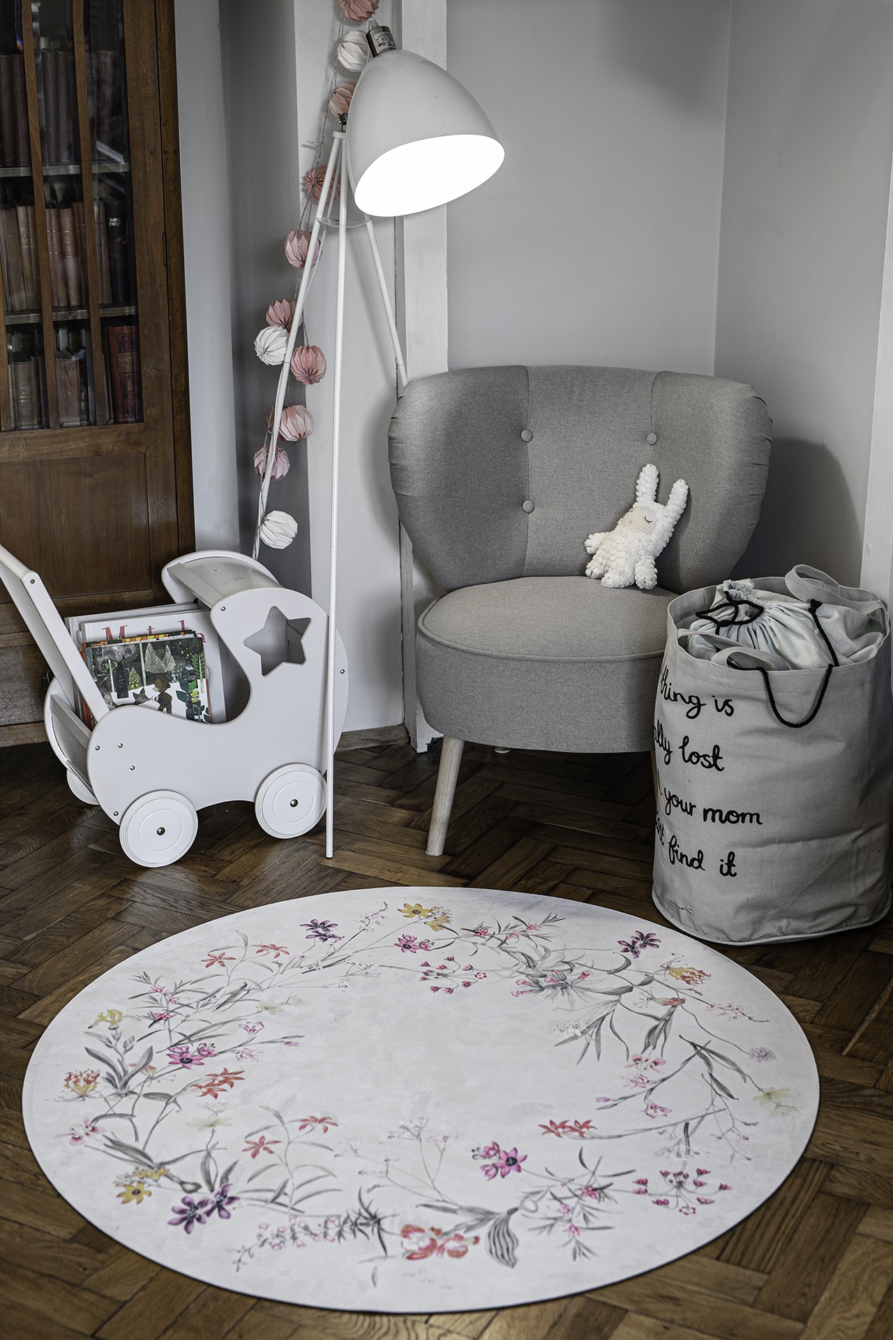 Tapete Redondo ECO de Bebé Jardim de Flores Canto de quarto com poltrona cinza, candeeiro de pé branco, carrinho de madeira branco e tapete floral redondo.
