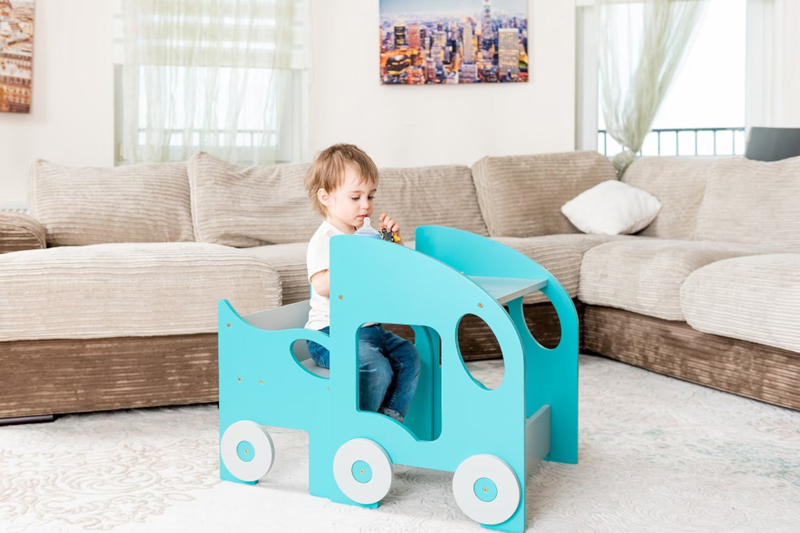 Mesa e Cadeira Infantil MONTESSORI Criança sentada em brinquedo de madeira azul claro em forma de carro numa sala com sofá castanho claro