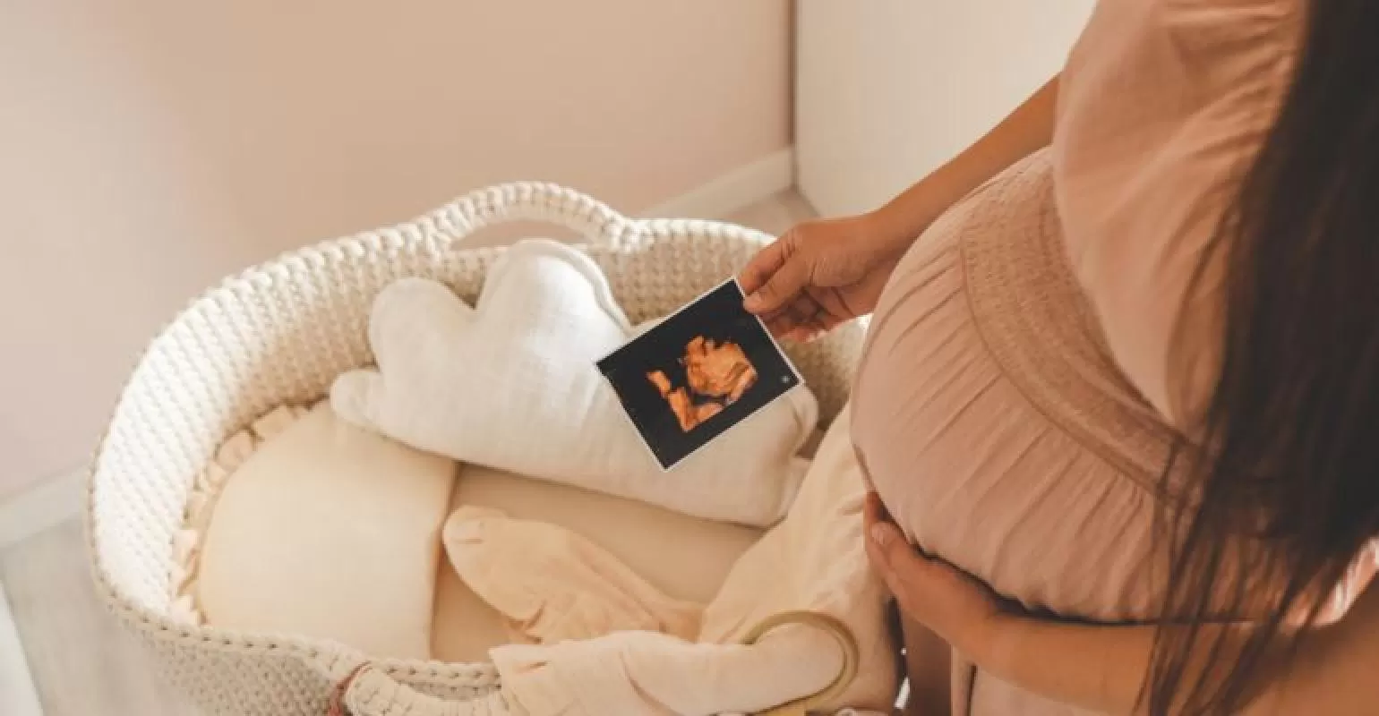 Berço branco de tecido com roupa de cama bege, travesseiro nuvem branca e mão de mulher grávida a segurar ecografia
