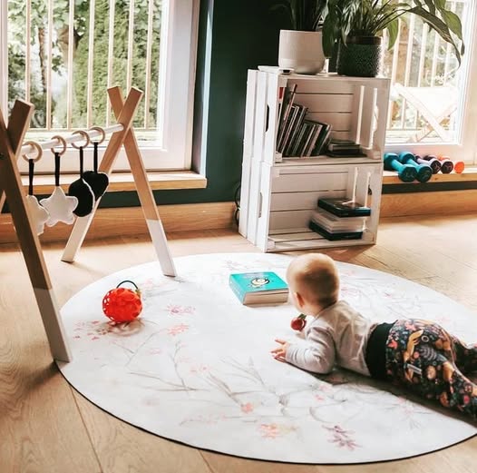 Tapete Redondo ECO de Bebé Jardim de Flores bebé deitado sobre tapete floral redondo com bolas e livros, mobiliário branco e plantas junto a janela
