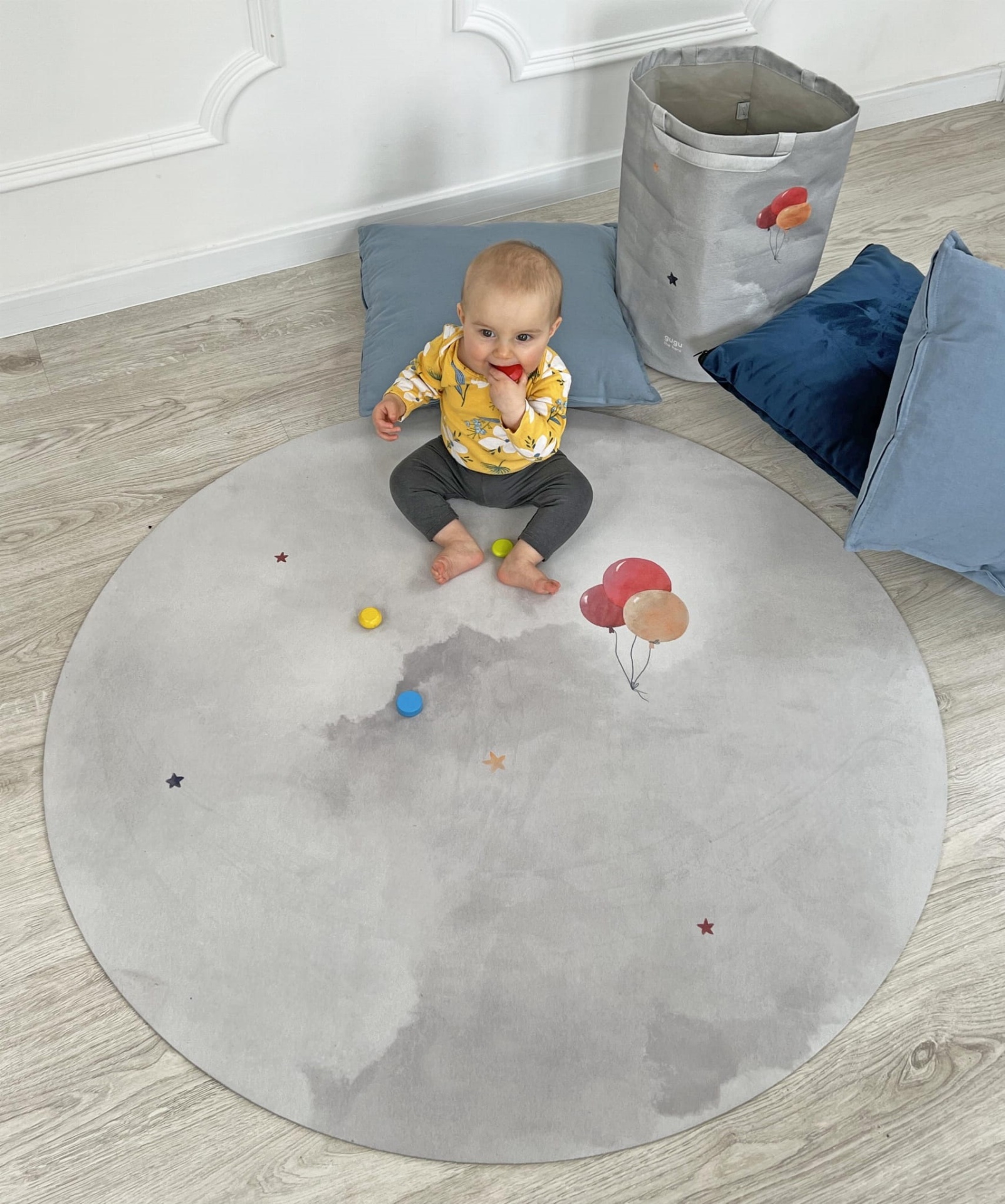 Tapete Redondo de Borracha Natural Balões PRIME Tapete redondo cinzento com padrão nuvem e balões, bebé sentado no centro, almofadas e saco de tecido ao lado